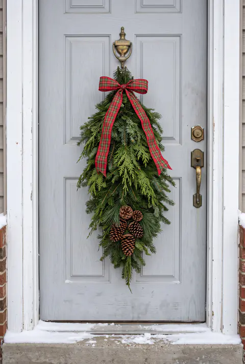 Long teardrop swag of fir, cedar and pine with a plaid ribbon and pinecones hanging from the knocker of a light gray door