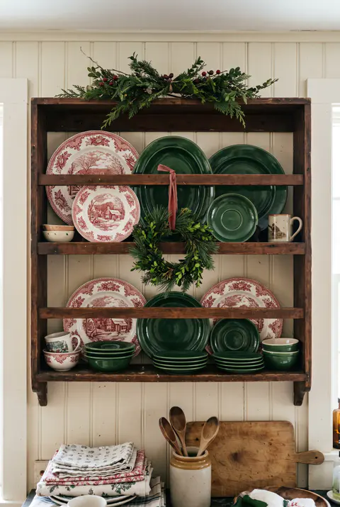 Wooden plate rack on a kitchen wall displaying red and white transferware Christmas plates and green dishes with a small garland on top