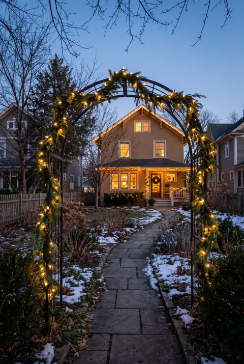 Garden path in a front yard with a metal arch wrapped in fir garland and warm white lights and a lit house in the background