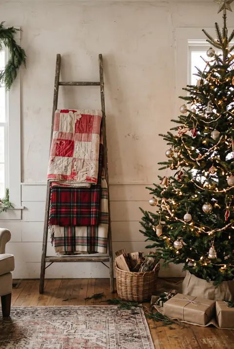 Vintage red and white patchwork quilt and wool plaid blankets folded over a wooden ladder next to a Christmas tree