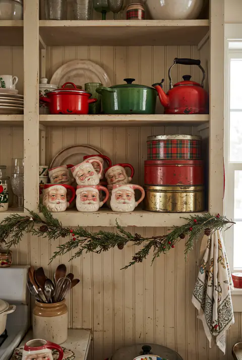 Open kitchen shelf with vintage-style Santa face mugs, red and green enamel cookware and plain cookie tins with a small garland