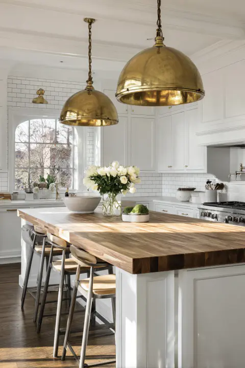 A bright and airy modern farmhouse kitchen showcasing two oversized antique brass dome pendant lights above a warm butcher block island. This kitchen ceiling lights idea provides both style and function, adding a touch of vintage warmth to the clean, white cabinet design.