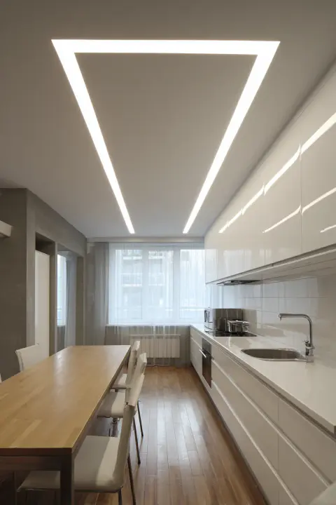 Kitchen Ceiling Lights, Bright and Functional with a triangle shape of led lights. Modern kitchen interior design. Open concept kitchen with white glossy cabinets, a long wooden dining table and a white countertop with a sink and stove. Recessed lighting. Minimalist style kitchen.