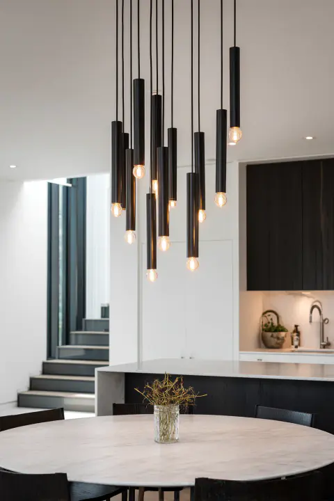 Modern kitchen ceiling lights featuring a striking cluster of black industrial mini pendants with Edison bulbs, arranged at various heights to form a sculptural centerpiece above a minimalist marble table.