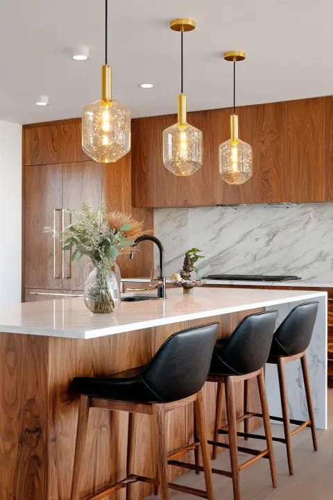 Modern kitchen with warm wood cabinetry, white marble countertops, and three brass and glass pendant lights over a kitchen island with black leather bar stools. Contemporary kitchen design, luxury home interiors, stylish kitchen decor
