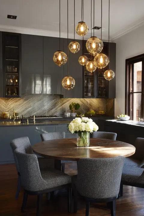 "Modern kitchen and dining area featuring dark grey cabinetry, a round wooden dining table with grey upholstered chairs, and a cluster of contemporary glass pendant lights. Marble backsplash and integrated appliances complete this elegant, high-end interior design.