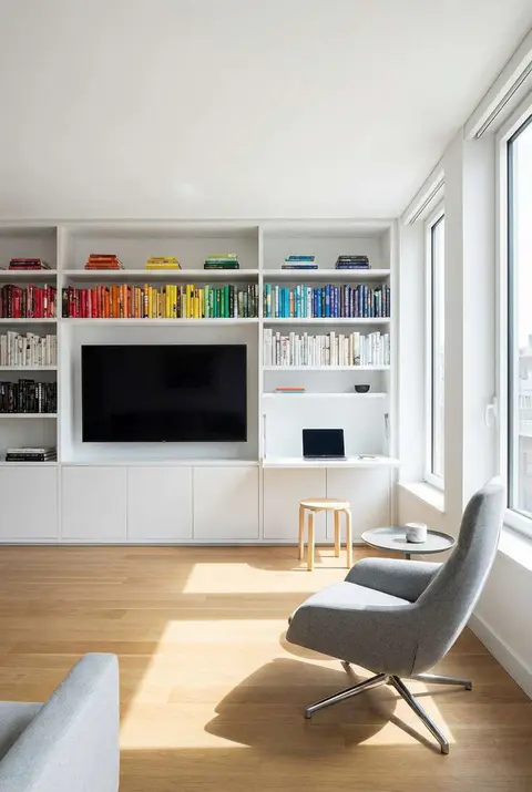 Bright, modern living room with white built-in wall unit featuring shelves of rainbow-colored books, a centered black television, and a hidden pull-down desk with a laptop and wooden stool. A light gray, modern swivel armchair sits on the light wood floor in the foreground, next to large windows letting in sunlight. Keywords: modern interior design, white built-in shelving, rainbow books, living room, light wood floor, gray armchair, natural light