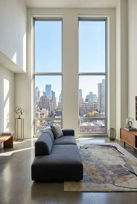 Modern luxury loft living room featuring soaring floor-to-ceiling windows with a bright city skyline view, a sleek grey sofa, and an abstract area rug on polished concrete floors.