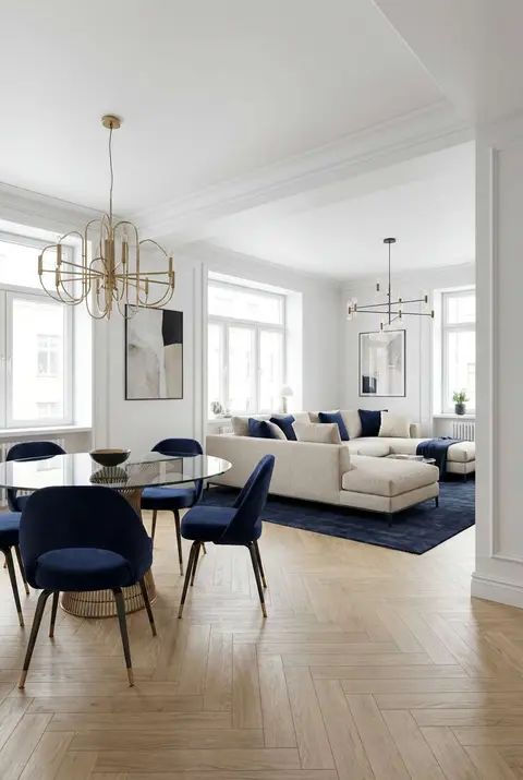 Elegant, light-filled living and dining area with white walls and large windows. The dining area features a round glass table with a woven base and six deep navy blue velvet chairs, illuminated by a modern gold geometric chandelier. The living area has a large, light beige sectional sofa on a dark blue area rug, with a modern black linear chandelier overhead. Keywords: bright interior, white walls, navy blue chairs, beige sectional sofa, gold chandelier, open concept living, herringbone wood floor