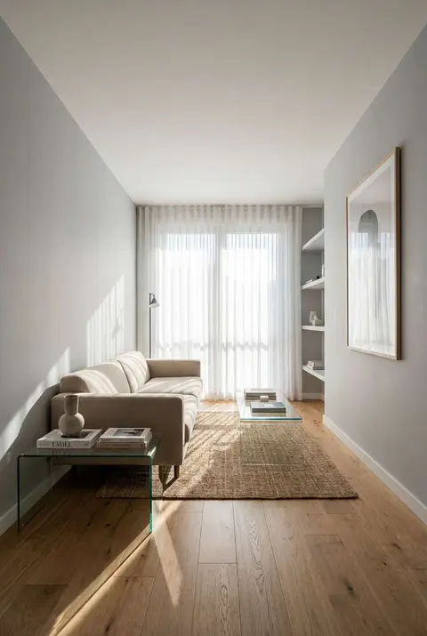 Bright minimalist living room bathed in natural sunlight through floor-to-ceiling sheer white curtains. The airy space features a modern beige sofa, a glass coffee table on a woven rug, light oak wood flooring, and large-scale contemporary wall art.