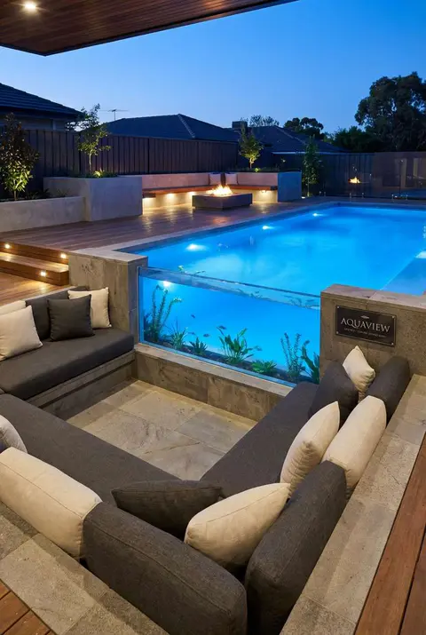 Luxury backyard oasis at dusk featuring a rectangular swimming pool with a unique glass-walled section showcasing an underwater aquarium or fish tank, adjacent to a sunken conversation pit with dark gray sectional seating and cream throw pillows, with a fire pit visible in the background. Modern pool design, sunken seating area, aquarium pool, outdoor entertaining