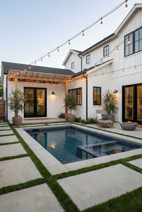 Modern farmhouse backyard with a rectangular swimming pool featuring dark blue tile, surrounded by large square concrete pavers separated by strips of green grass, leading up to a white board-and-batten style house with black-trimmed windows and glass doors, all illuminated by overhead string lights at twilight. Farmhouse style, concrete paver patio, backyard pool design, outdoor lighting