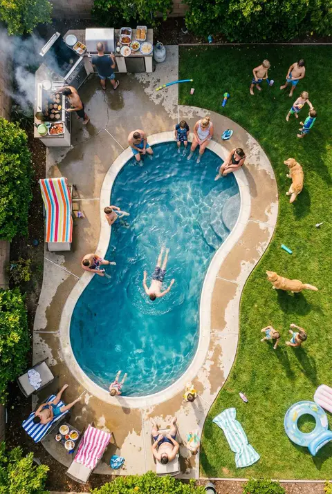 Aerial view of a lively backyard pool party on a sunny day. People are swimming, lounging on the deck chairs, and socializing around a kidney-shaped pool. A barbecue grill is actively smoking near the top, and several children and two dogs are playing on the surrounding green lawn. Keywords: aerial pool party, backyard gathering, kidney shaped pool, summer fun, BBQ, family activity.