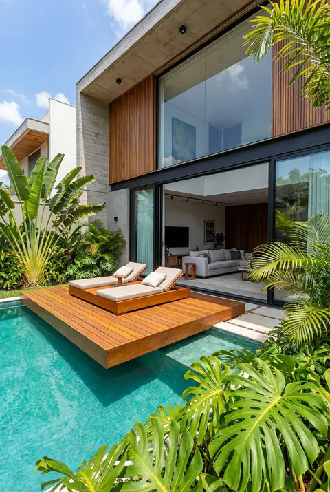 Luxurious modern home with a swimming pool integrated with tropical landscaping. A square wooden deck floats partially over the turquoise pool water, holding two modern chaise lounge chairs. The house features large glass sliding doors, wood paneling, and concrete accents, surrounded by large-leaf tropical plants like Monstera and banana trees. Modern tropical architecture, floating deck, integrated pool design, indoor-outdoor living