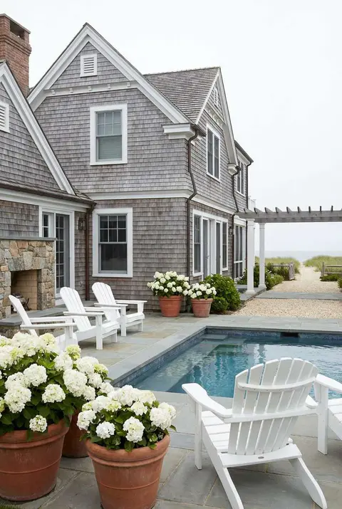 Coastal or Hamptons style backyard featuring a rectangular swimming pool with blue tile trim, set next to a shingled gray cedar house with white trim and a stone fireplace. The patio area has large blue-gray flagstones, white Adirondack chairs, and large terracotta pots overflowing with white hydrangeas. Beach house, New England style, classic pool area, seaside garden.