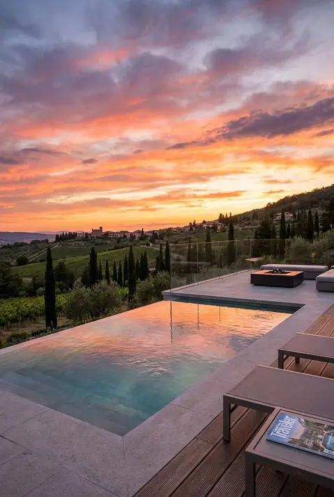 Stunning infinity-edge swimming pool overlooking rolling Tuscan hills and vineyards at sunset. The sky is filled with vibrant orange and pink clouds reflecting in the pool's surface. The pool deck features light stone coping, modern lounge chairs, and a glass safety railing, emphasizing the panoramic, picturesque landscape view.